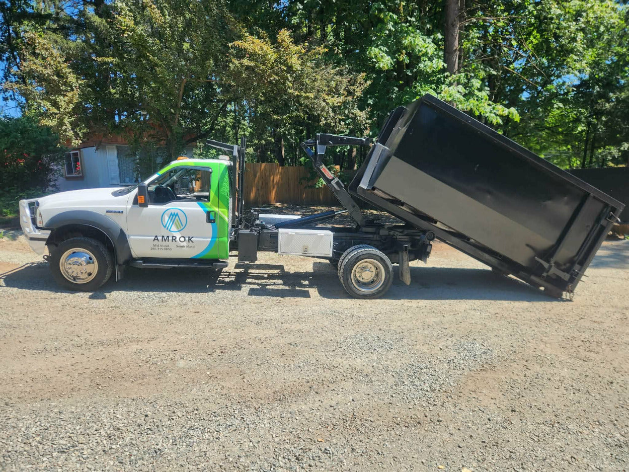A white utility truck with green and blue graphics and the brand name AMROK, towing a large black dump trailer that is tilted upward, parked on a gravel surface outdoors with trees in the background.